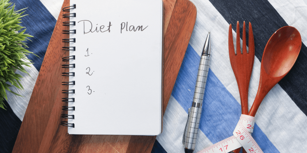 식탁 보 위 다이어트 계획이라고 적은 노트, 수저세트, 펜을 위에서 찍은 사진